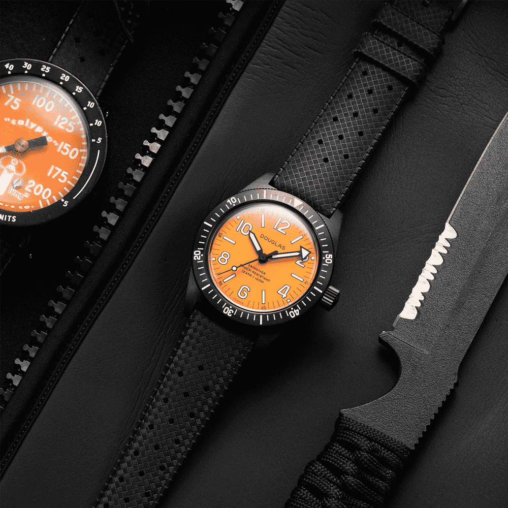 A black watch with an orange dial, black textured strap, and rotating bezel is displayed alongside a similar watch, a black case, and a black tactical knife with a serrated edge on a dark surface.